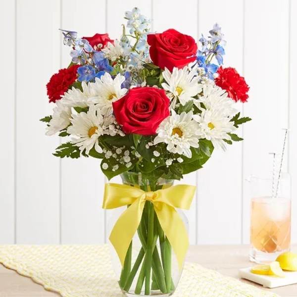 Bouquet of red roses, white daisies, and blue flowers in a glass vase with a yellow ribbon