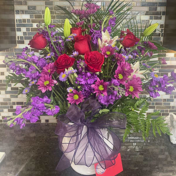Purple and red mixed flower arrangement in a white vase with a ribbon
