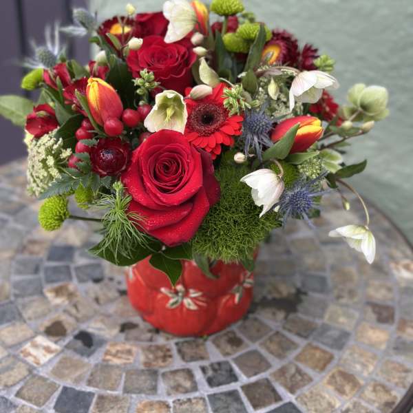 Compact arrangement of red roses, tulips, and mixed blooms in a red ceramic vase on a mosaic table.