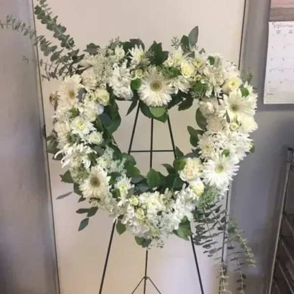 White floral wreath on a black stand with greenery accents