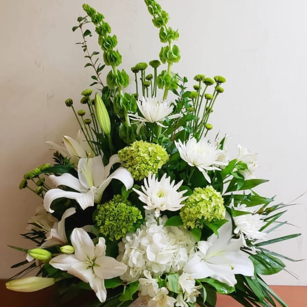 White and green floral arrangement with lilies, hydrangeas, mums, and bells of Ireland in a low white basket