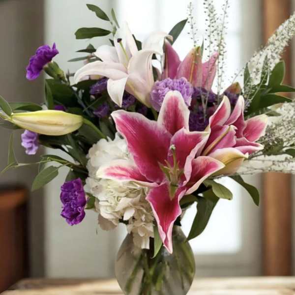 Pink lilies, white hydrangea, and purple flowers arranged in a clear glass vase on a wooden table