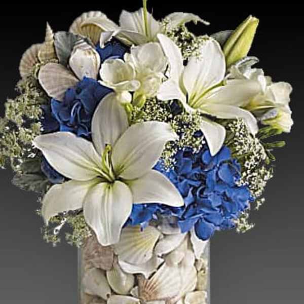 White lilies and blue hydrangeas in a shell-filled glass vase