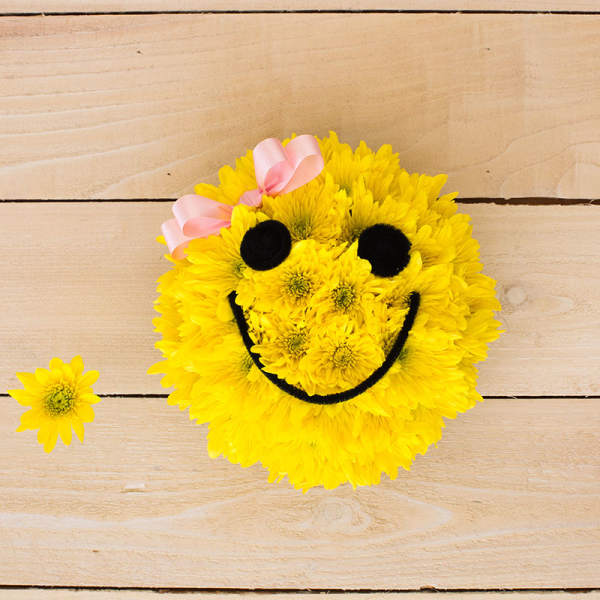 Round yellow floral arrangement shaped like a smiling face with black features and a pink bow.