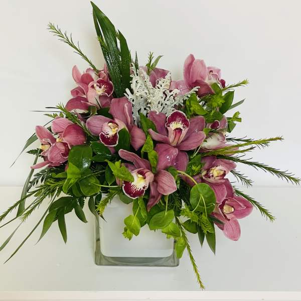 Pink orchid arrangement in a clear square vase with green foliage