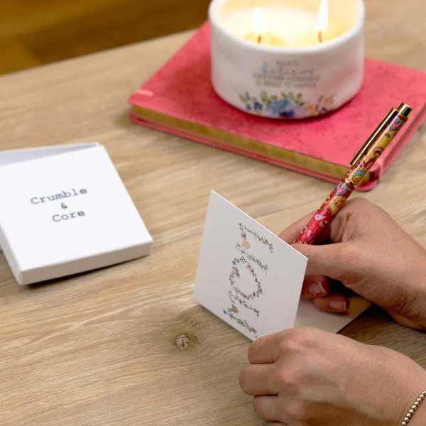Hands writing in a floral greeting card beside a lit candle and card box
