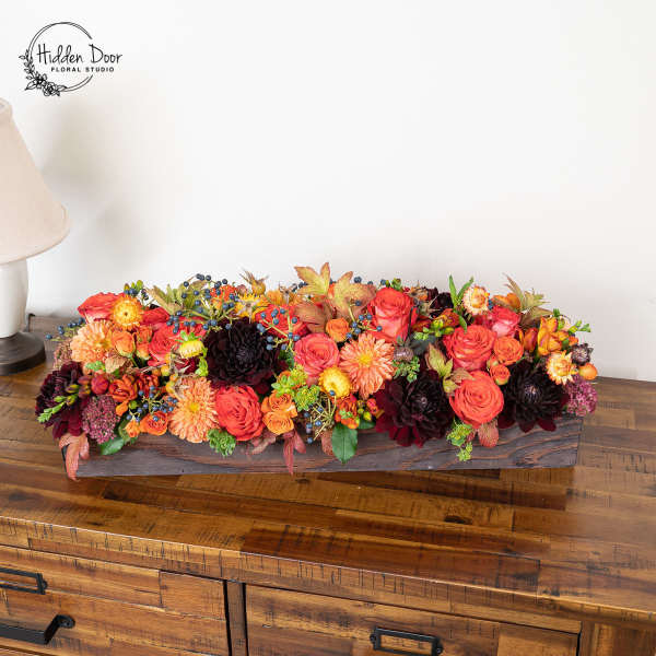 Long low floral arrangement with orange, red, and dark burgundy blooms in a wooden box