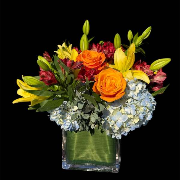 Orange roses and yellow lilies in a square glass vase with blue hydrangeas