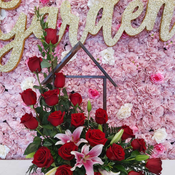 Red roses and lilies in a black house-shaped stand before a pink floral wall