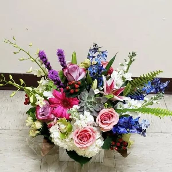Low mixed arrangement of pink roses, lilies, gerbera daisy, blue delphinium, orchids, and succulents.