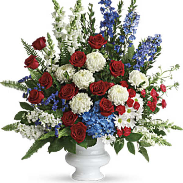 Large red, white, and blue flower arrangement with roses, mums, and delphinium in a white pedestal vase