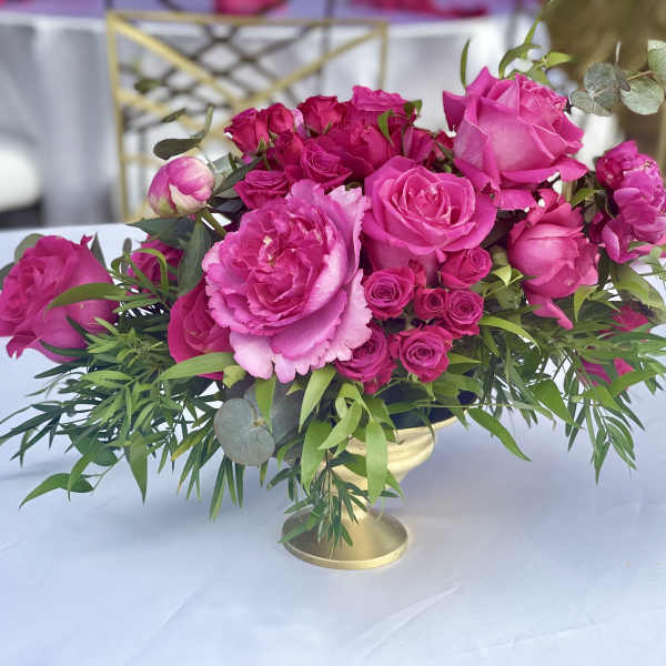 Low centerpiece of bright pink roses and peonies with greenery in a gold compote vase on a white table