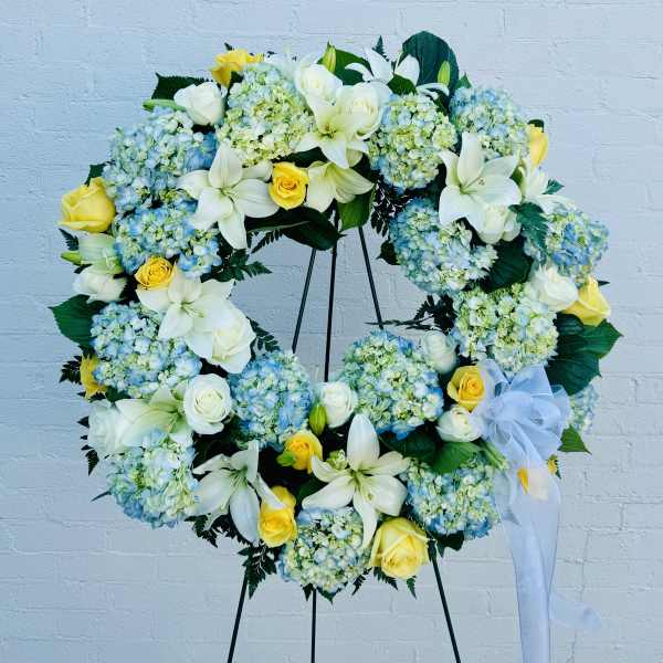 Large floral wreath with white lilies, yellow roses, and blue hydrangeas on a stand