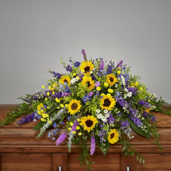 Bright casket spray of yellow sunflowers and mixed purple and white flowers on a wooden casket