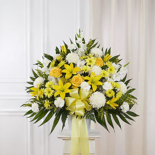 Large yellow and white sympathy arrangement with lilies, roses, and mums in a white urn with a yellow bow