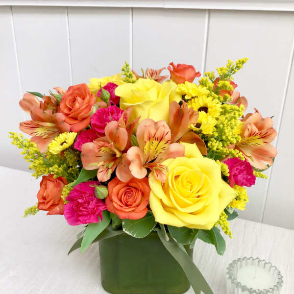 Bright arrangement of yellow roses, orange alstroemeria, and pink blooms in a green cube vase.