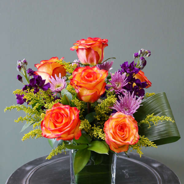 Bright arrangement of orange roses and purple flowers in a glass cube vase with a folded green leaf