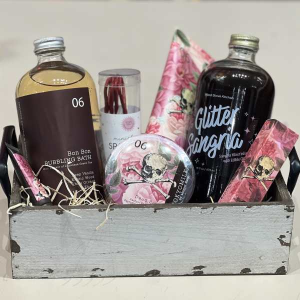 Wooden crate gift set with bath soak, sangria mixer, and pink floral skull-themed self-care items.