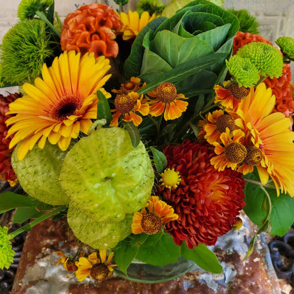 Bouquet of yellow gerbera daisies, orange mums, and green ornamental pods in a glass vase