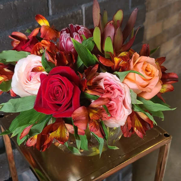 Low arrangement of red, pink, and peach roses with orange alstroemeria in a clear vase on a metal stand