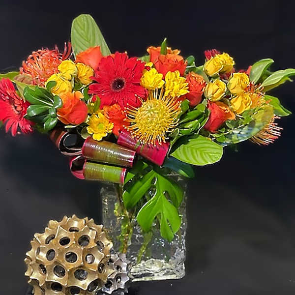 Bright red, orange, and yellow flower arrangement with tropical accents in a clear glass cube vase