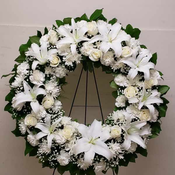 Large white floral wreath with lilies, roses, and mums displayed on a metal easel stand
