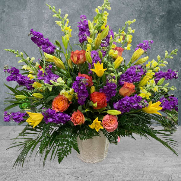Large arrangement of purple and yellow flowers with orange roses in a white vase