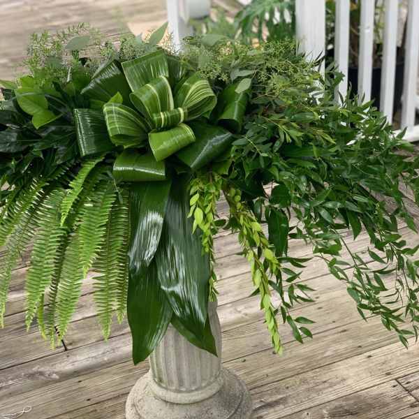Green foliage arrangement in a pedestal vase with a large bow