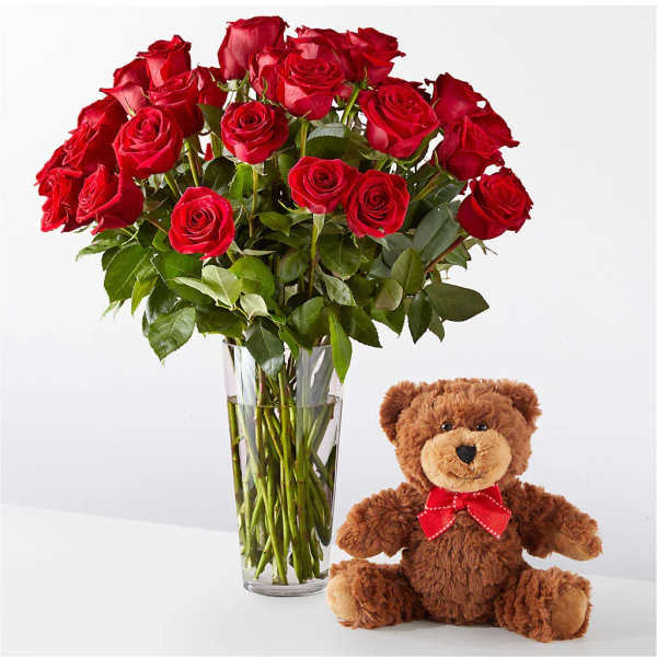 Red roses in a clear glass vase beside a teddy bear with a red bow
