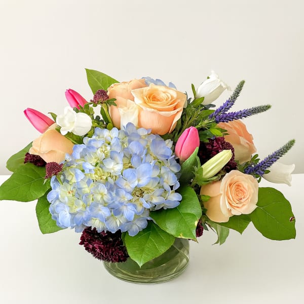 Bouquet of peach roses, blue hydrangea, and pink tulips in a glass vase