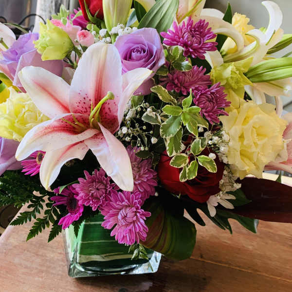 Mixed arrangement of pink lilies, purple roses, red roses, and yellow blooms in a square glass vase