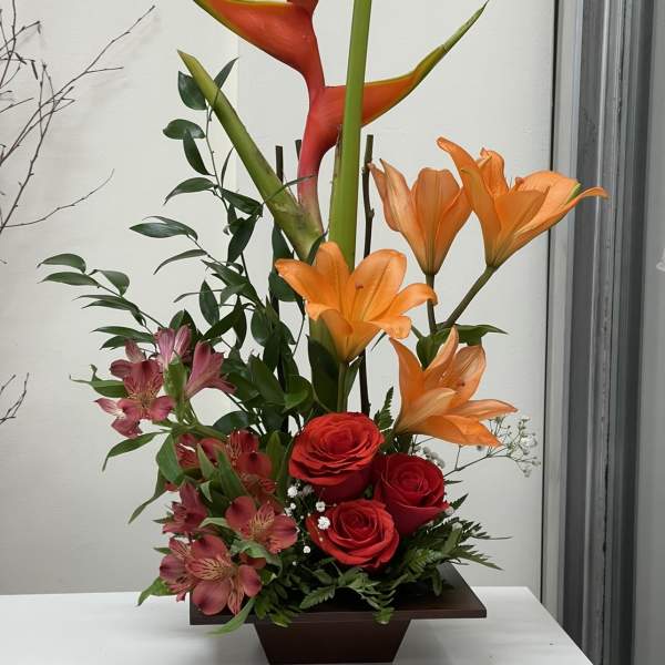 Tropical arrangement with orange heliconia, red roses, and pink alstroemeria in a dark vase