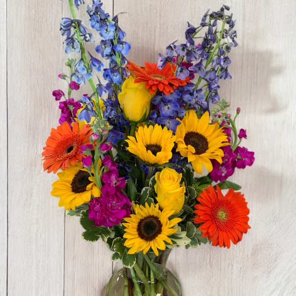 Bright mixed bouquet with sunflowers, gerberas, roses, and blue delphinium in a glass vase