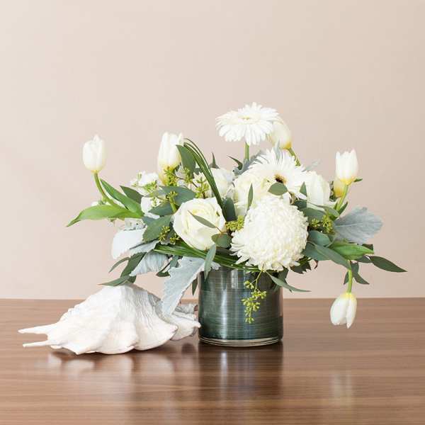 Low arrangement of white flowers in a glass vase beside a large white seashell on a wooden surface.