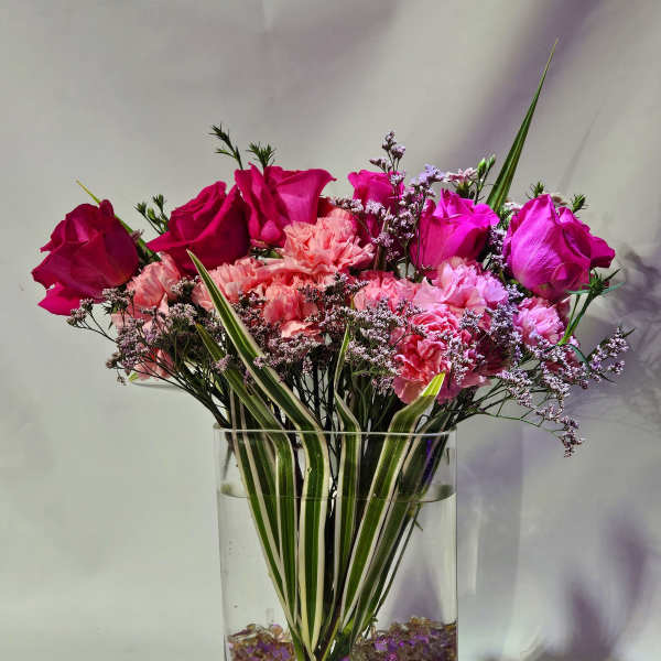 Tall arrangement of hot pink roses and pink carnations in a clear rectangular vase with striped foliage.