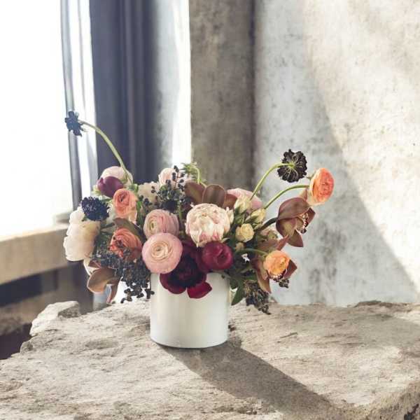 Low white vase with mixed pink, peach, and burgundy flowers on a stone surface