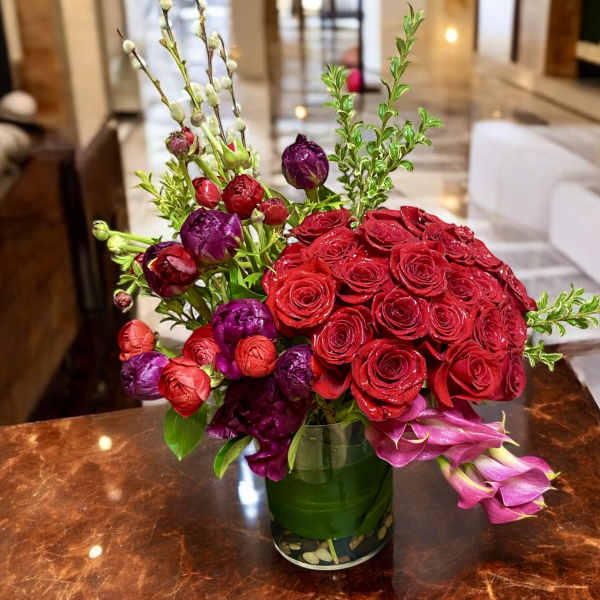 Lush red roses with purple and red blooms and pink calla lilies in a glass cylinder vase.