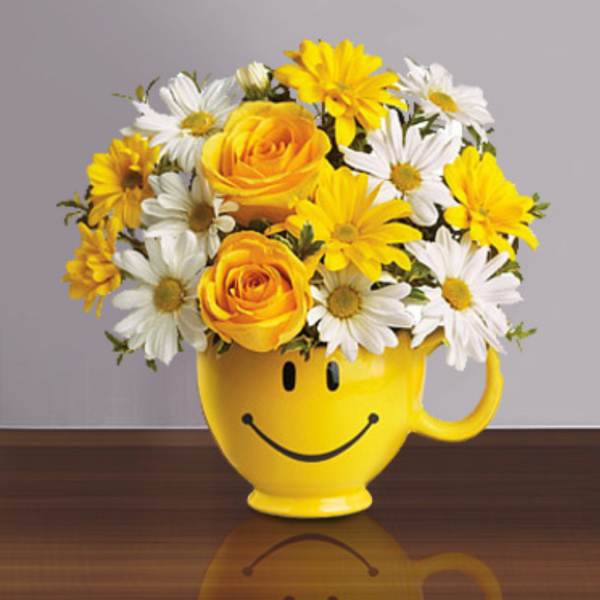 Yellow roses and white and yellow daisies arranged in a yellow smiley-face mug on a table