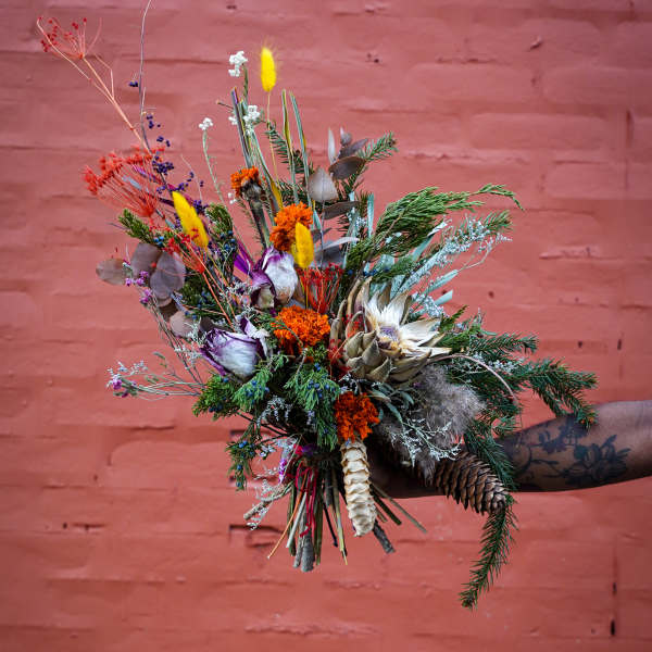 Wildflower bouquet with dried blooms and pinecones held against a pink brick wall
