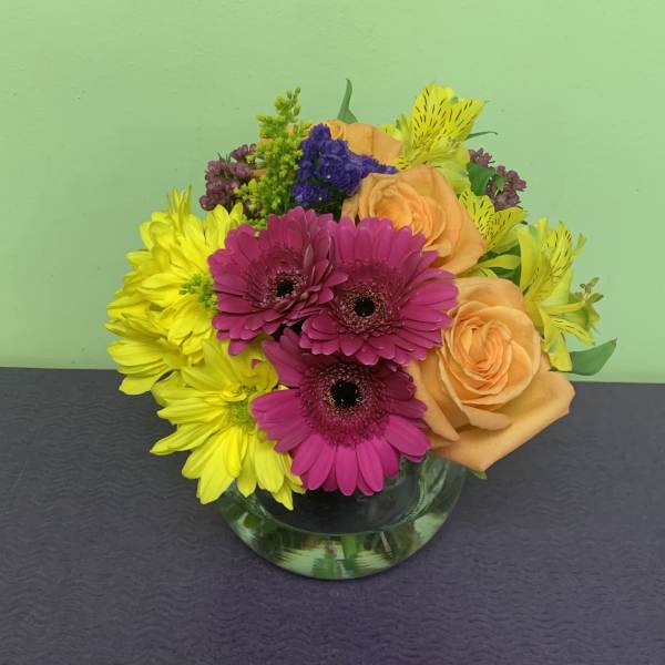 Mixed bouquet of pink gerbera daisies, yellow blooms, and peach roses in a glass vase