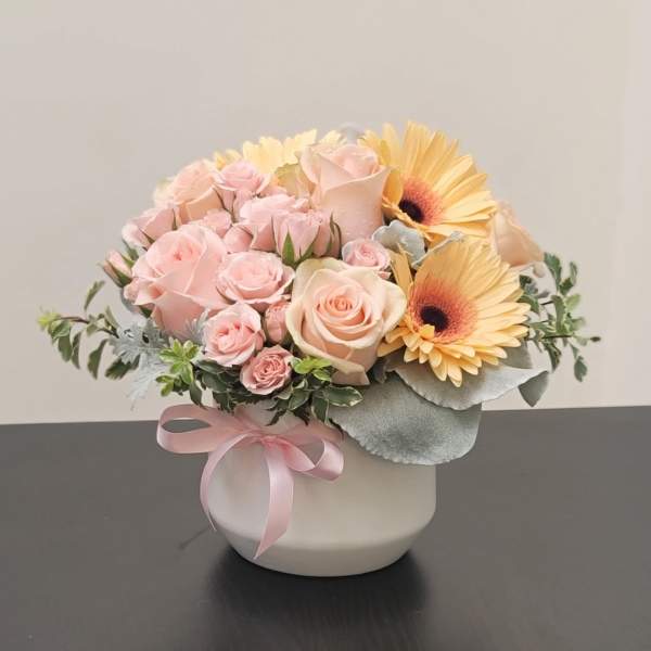 Pink roses and yellow gerbera daisies in a white vase with a pink ribbon