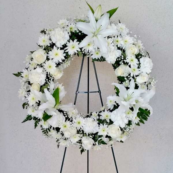 Large white floral wreath with lilies, roses, and daisies on a metal easel stand.