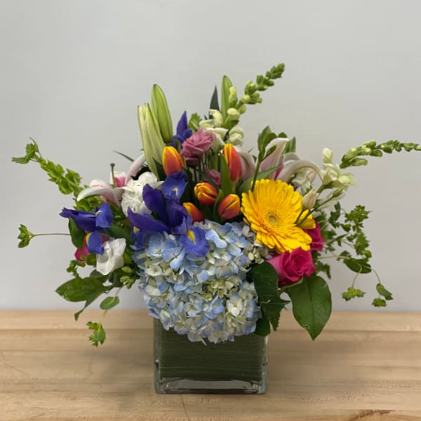 Mixed bouquet in a square glass vase with blue hydrangeas, yellow gerbera, and tulips