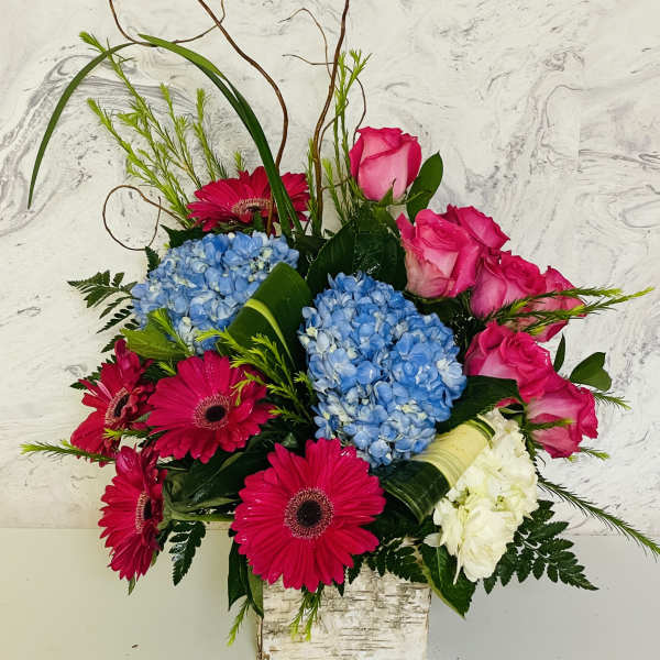 Bright bouquet of pink roses, blue hydrangeas, and magenta gerbera daisies in a square vase