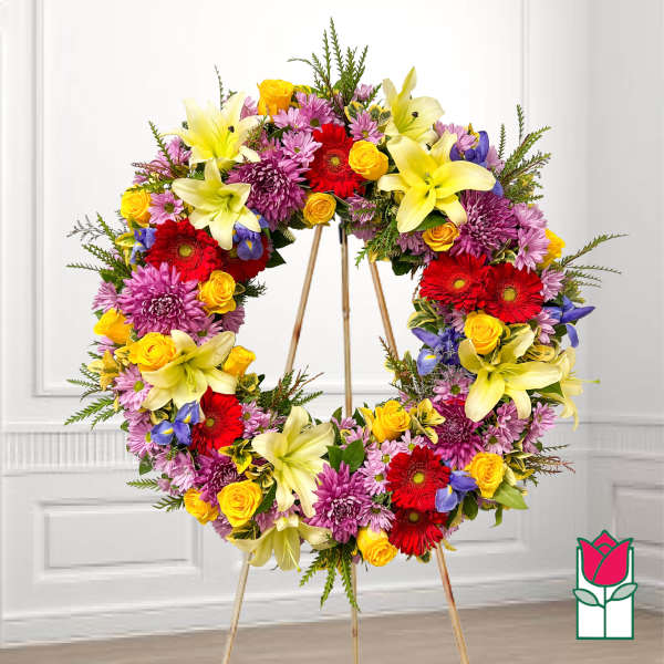 Large floral wreath on a stand with yellow lilies, red gerberas, and purple blooms