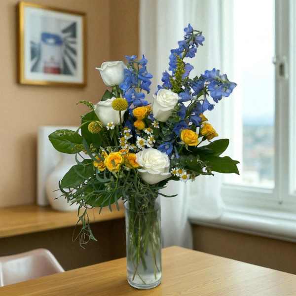 Tall arrangement of white roses, blue delphinium, and yellow blooms in a clear glass vase on a wooden table