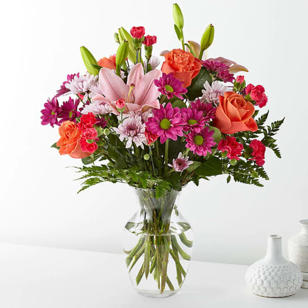 Mixed bouquet of pink lilies, orange roses, and magenta daisies in a clear glass vase