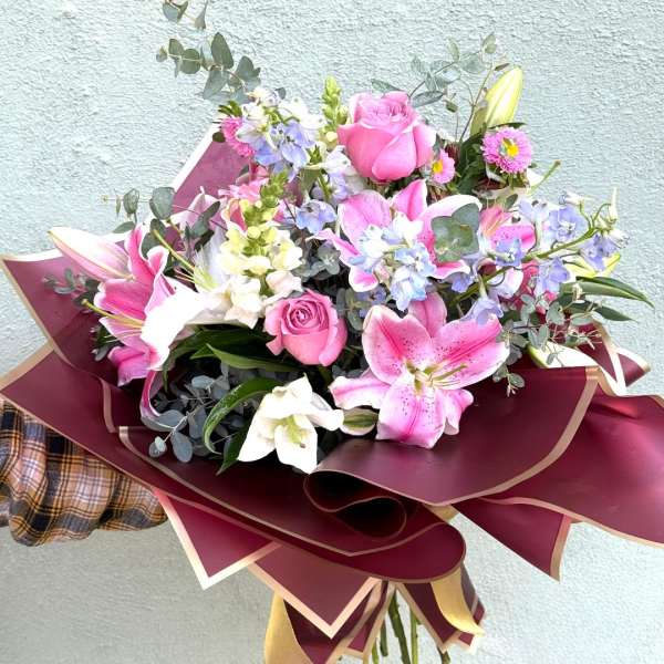 Hand-tied bouquet of pink roses, lilies, and mixed blooms wrapped in burgundy and gold paper.