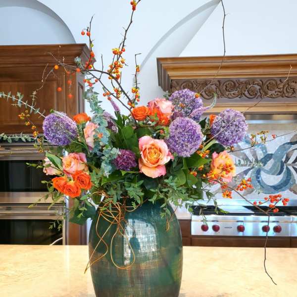 Tall floral arrangement with roses, purple allium, and berry branches in a green vase