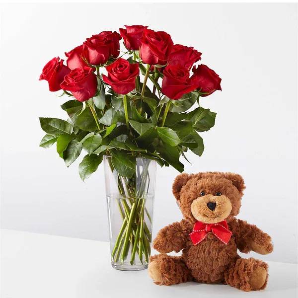 Red roses in a clear vase beside a teddy bear with a red bow